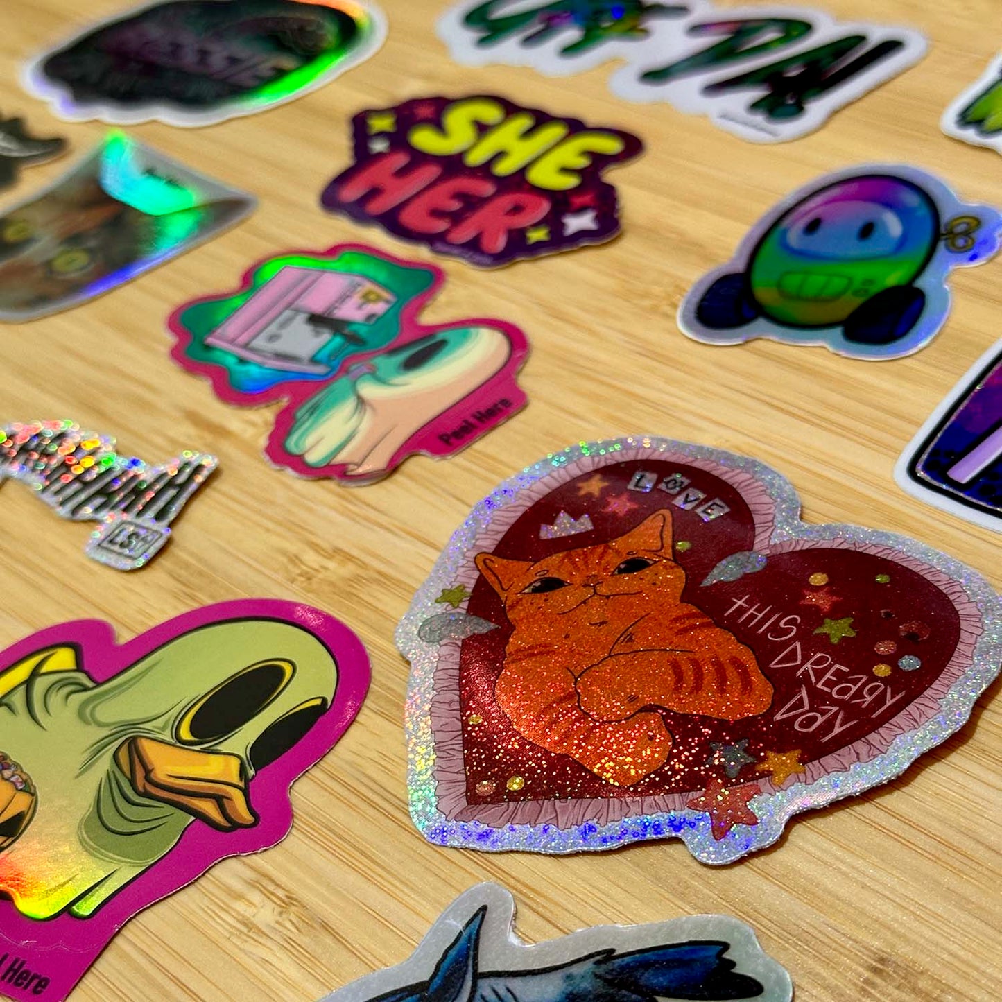 A wood surface featuring holographic stickers. The sticker in focus in a glitter holographic sticker with a cat in a valentine and text reading "this dreary day"