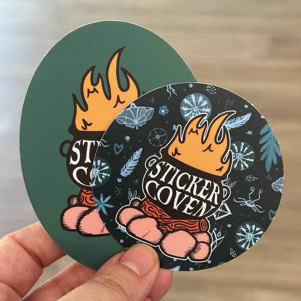 A hand holding a circle sticker and an oval sticker with graphics of a cauldron on a campfire with text "Sticker Coven"