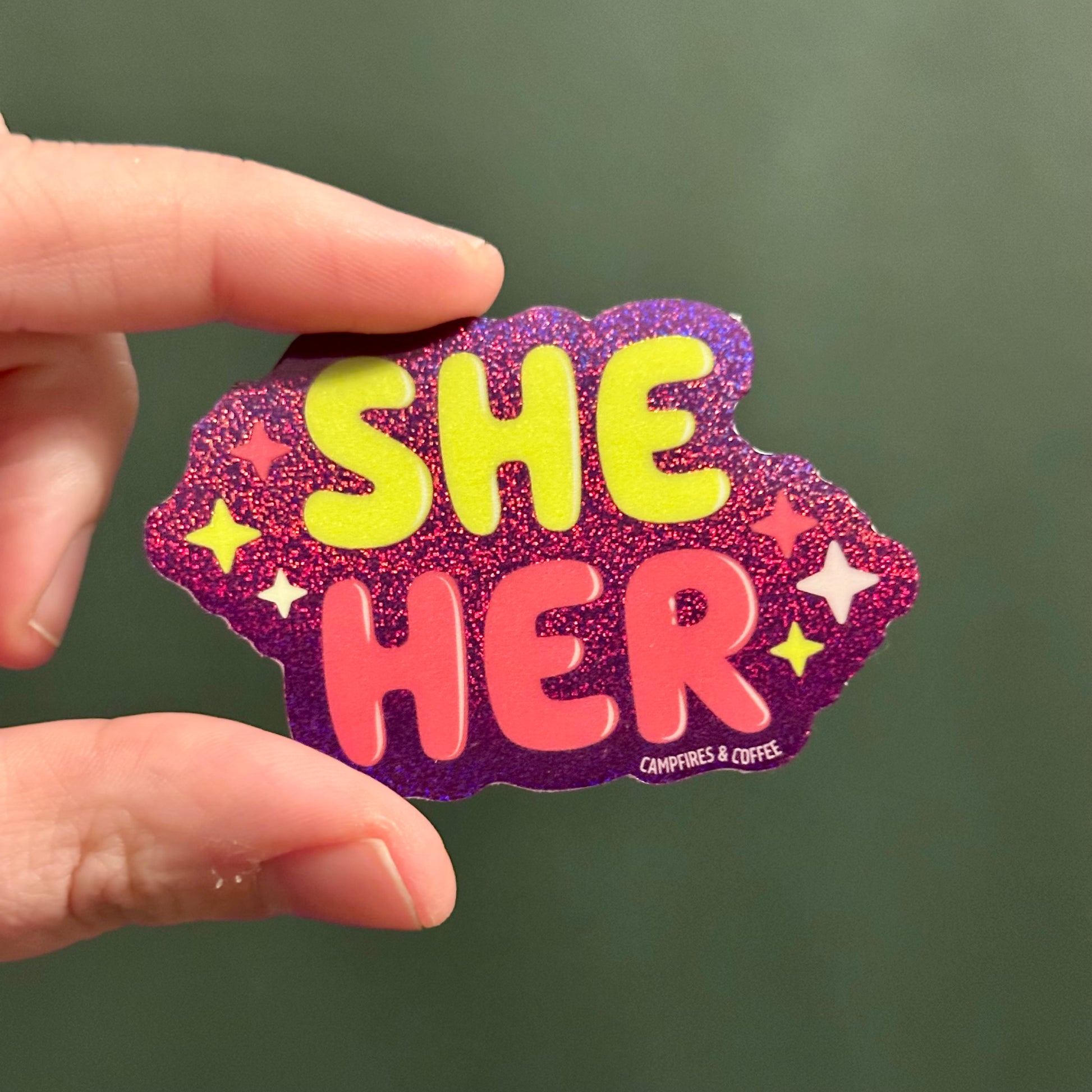 purple glitter holographic sticker with the text "She Her" in green and pink font