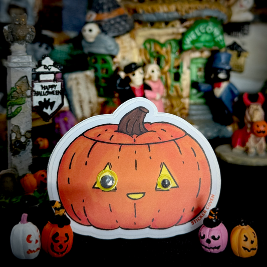 A cute cartoon jack-o-lantern sticker with a smiling face and googly eyes, displayed in a themed Halloween setting with small pumpkins and festive decorations.
