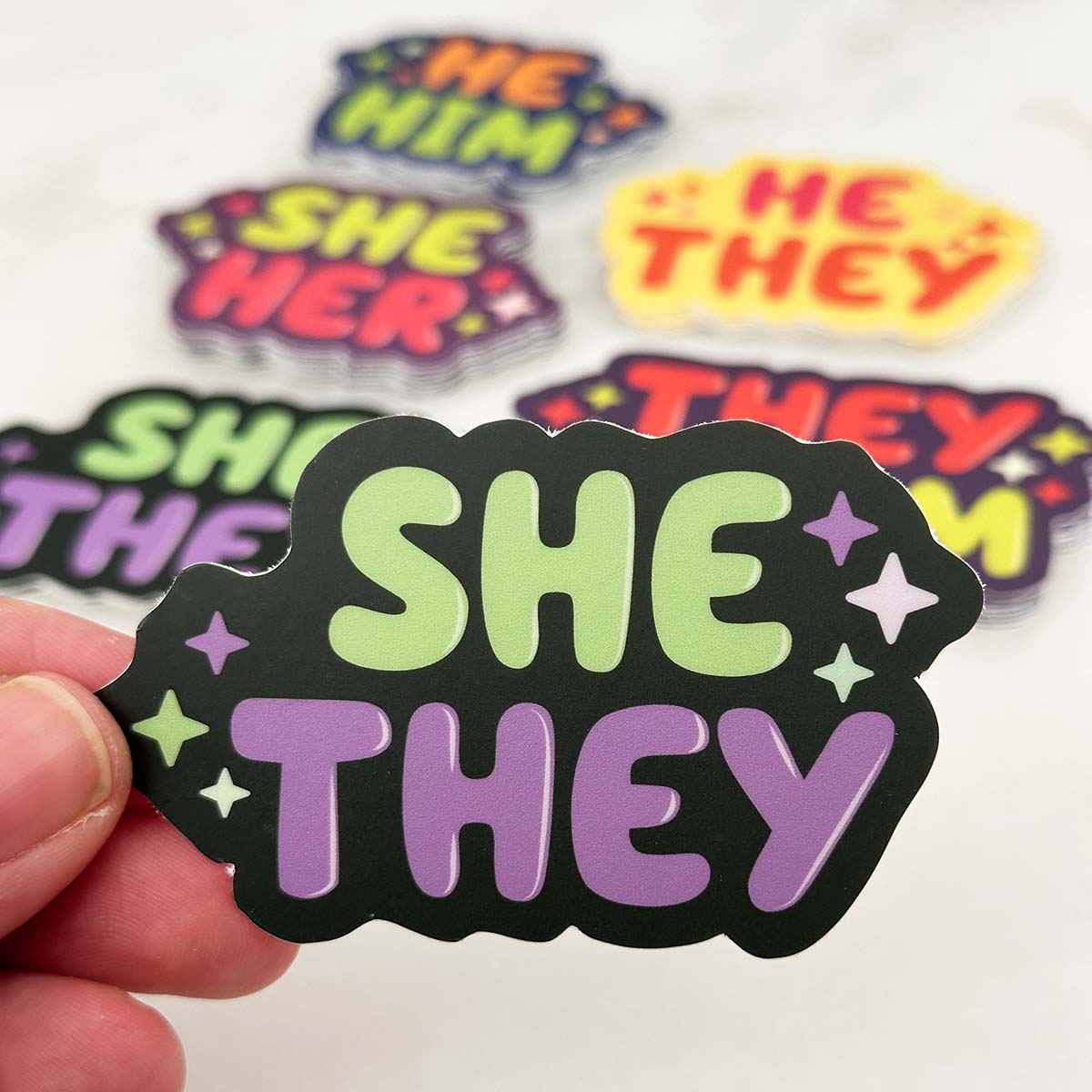 In the foreground: die cut sticker with text "she they" decorated with stars. In the background: an assortment of pronoun stickers in stacks on a white table