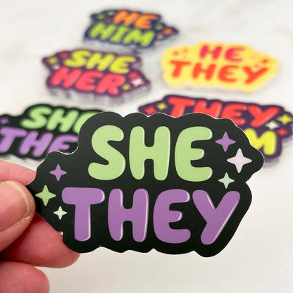 In the foreground: die cut sticker with text "she they" decorated with stars. In the background: an assortment of pronoun stickers in stacks on a white table