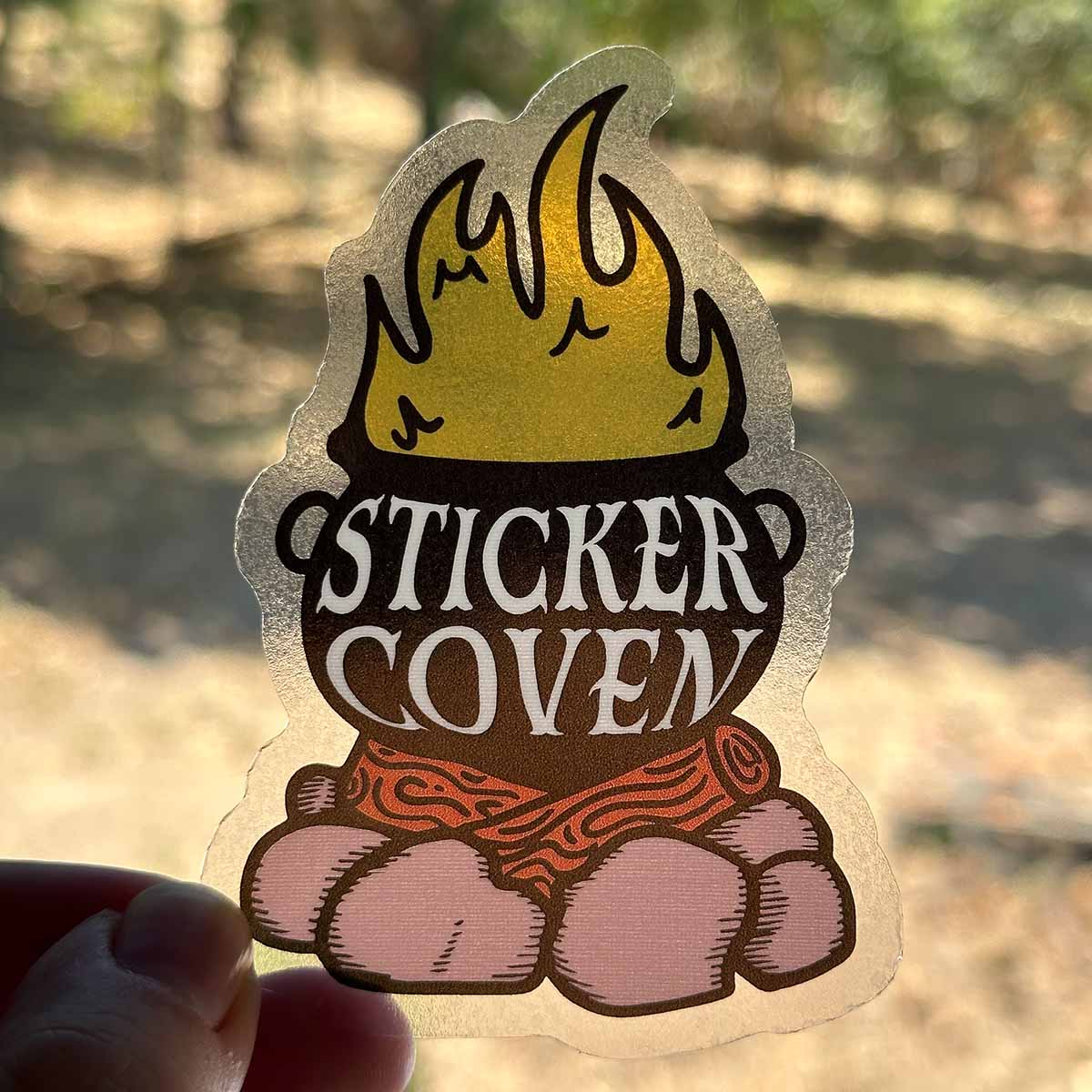 A hand holding a clear die cut sticker up to a window to highlight transparency. The sticker features a cauldron on a campfire with white text "Sticker Coven"