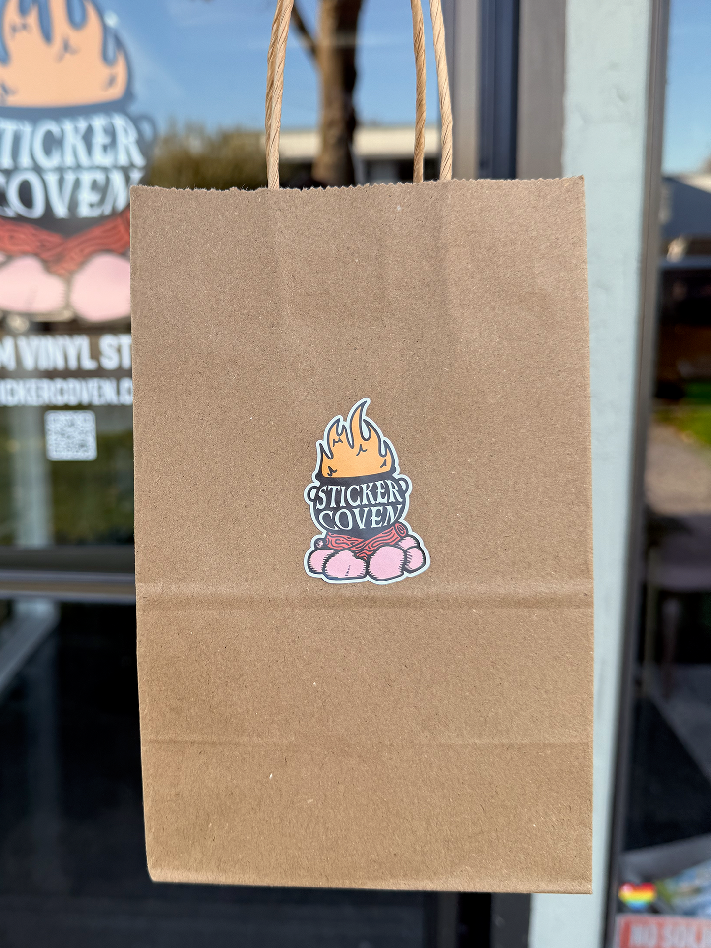 Brown paper bag with a 'Sticker Coven' logo economy sticker in front of a store window.