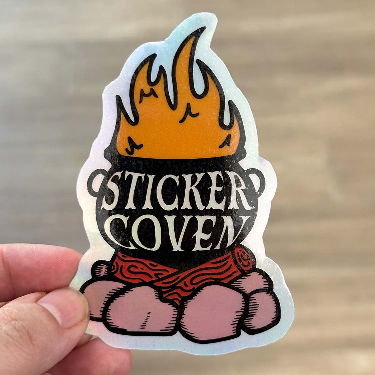 A hand holding a die cut cauldron on a campfire holographic sticker with glossy laminate. Text on the sticker reads "Sticker Cauldron"