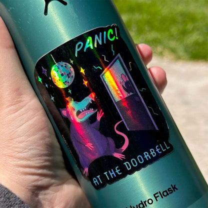 A hand holding a green water bottle with a holographic sticker. The sticker depicts a possum panicking at the doorbell with a mirror ball overhead. Text reads: "Panic! at the doorbell"