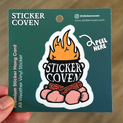 A hand holding a green hanging card with a cauldron on a campfire sticker and the text "Sticker Coven"