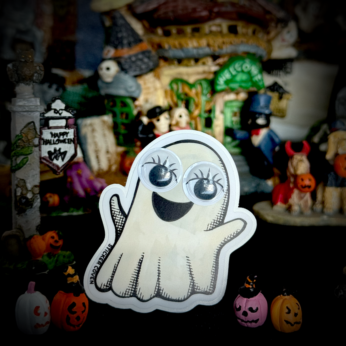 A cute cartoon ghost sticker with large googly eyes and a smiling mouth, displayed in front of a Halloween themed miniature village scene with pumpkins and figurines.