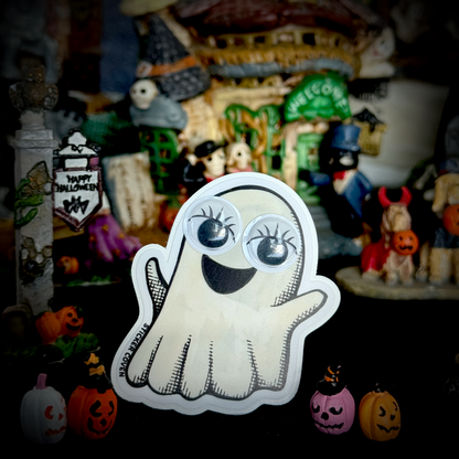 A cute cartoon ghost sticker with large googly eyes and a smiling mouth, displayed in front of a Halloween themed miniature village scene with pumpkins and figurines.