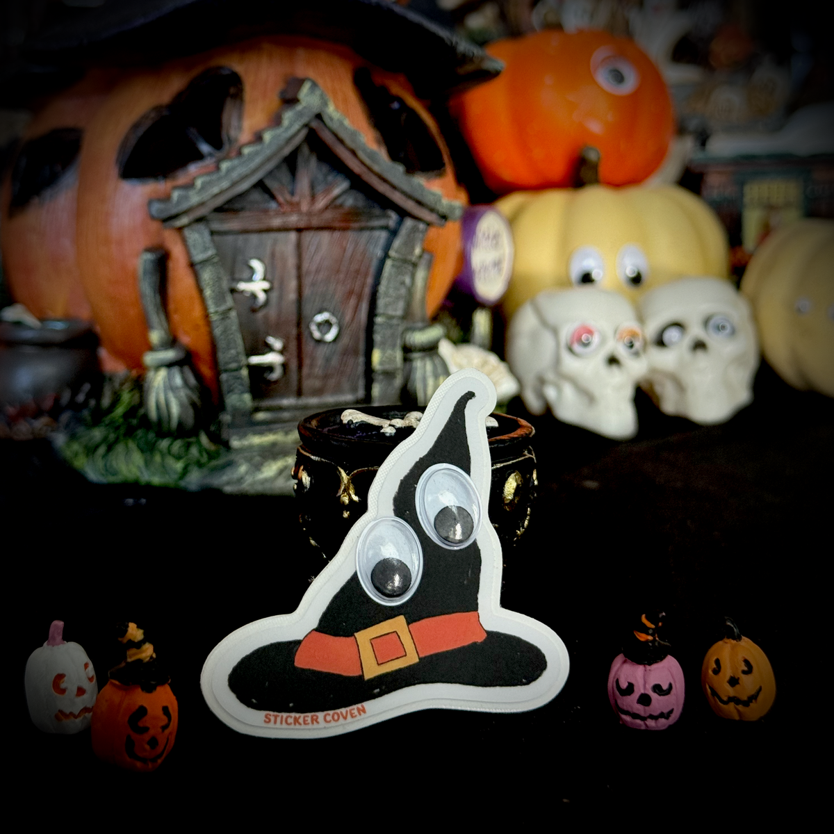 A black witch hat sticker with googly eyes, a red-orange belt, and the text "Sticker Coven" at the bottom. The sticker is displayed in the foreground on a dark surface, with small jack-o-lanterns and a  pumpkin house, pumpkins, and skulls blurred in the background.